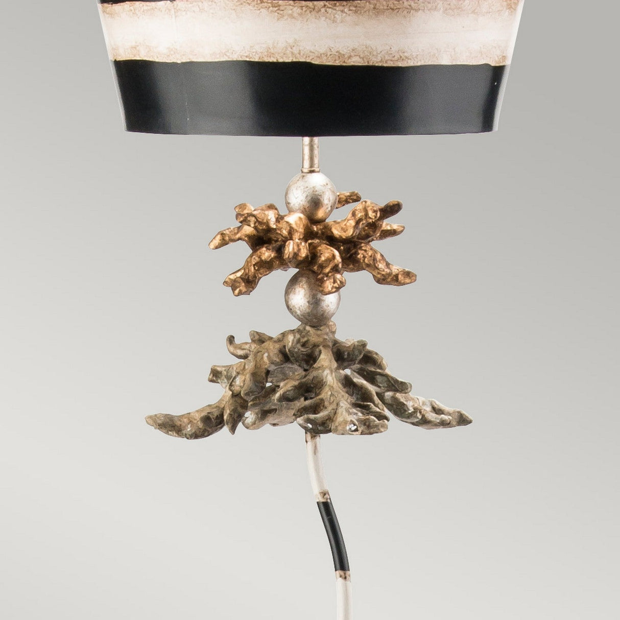 A close-up of the South Beach 1 Light - Black, Putty & Gold Leaf highlights its tropical elegance with a metallic ornamental base reminiscent of twisted branches or coral. The lamp is adorned with a cylindrical, black and white striped shade set against a simple gray backdrop.