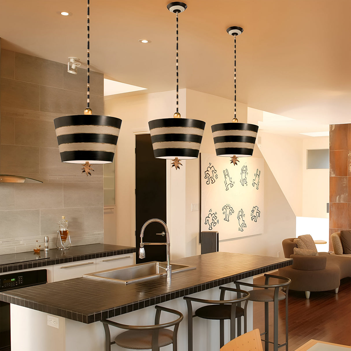 Modern kitchen with a large island countertop featuring the South Beach 1 Light Pendant in Black, Putty, and Gold Leaf. Wooden chairs surround the island. In the background, a cozy seating area and a wall with decorative figures add coastal elegance, while natural light beautifully illuminates the space.