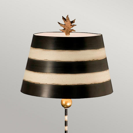 The South Beach 1 Light Table Lamp in Black, Putty, and Gold Leaf exudes coastal charm with its dark shade featuring horizontal cream stripes and a leaf-shaped finial. Its slender base is elegantly accented by a Gold Leaf orb, all set against a plain gray backdrop.