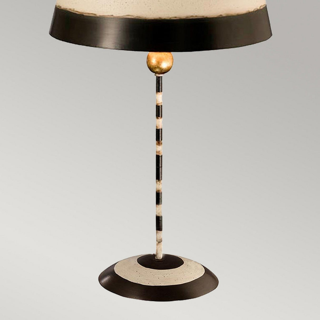 The South Beach 1 Light Table Lamp in Black, Putty & Gold Leaf exudes coastal charm with its vintage design and a round beige and black shade. The striped-patterned pole is highlighted by a gold accent near the top, while its circular base complements the cohesive color scheme against a simple gray backdrop.