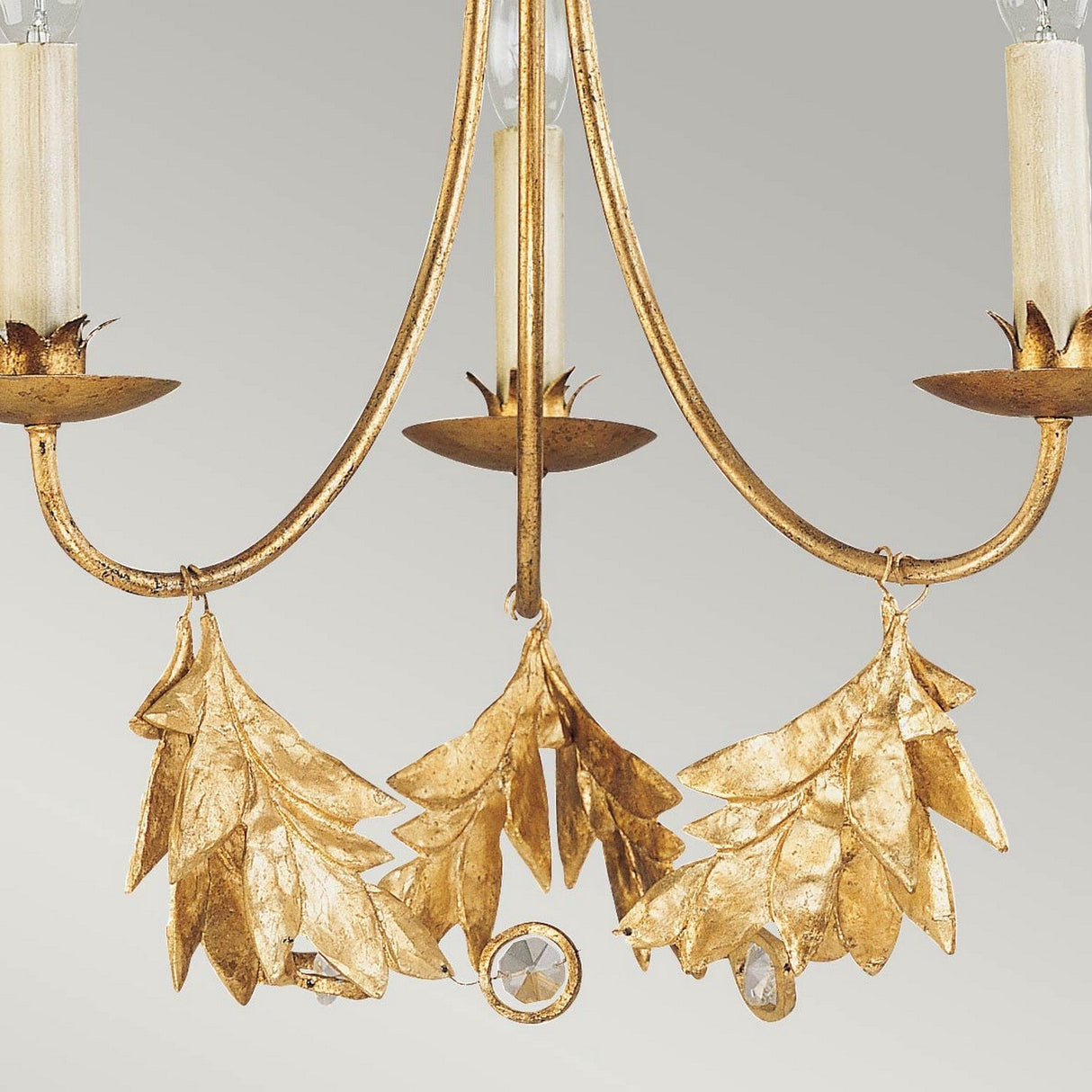 A close-up of the Sweet Olive 3 Light Chandelier in gilded gold reveals its intricate leaf accents, featuring two candle-shaped lights and decorative gold leaves elegantly hanging beneath a shimmering crystal medallion, all set against a plain gray background.