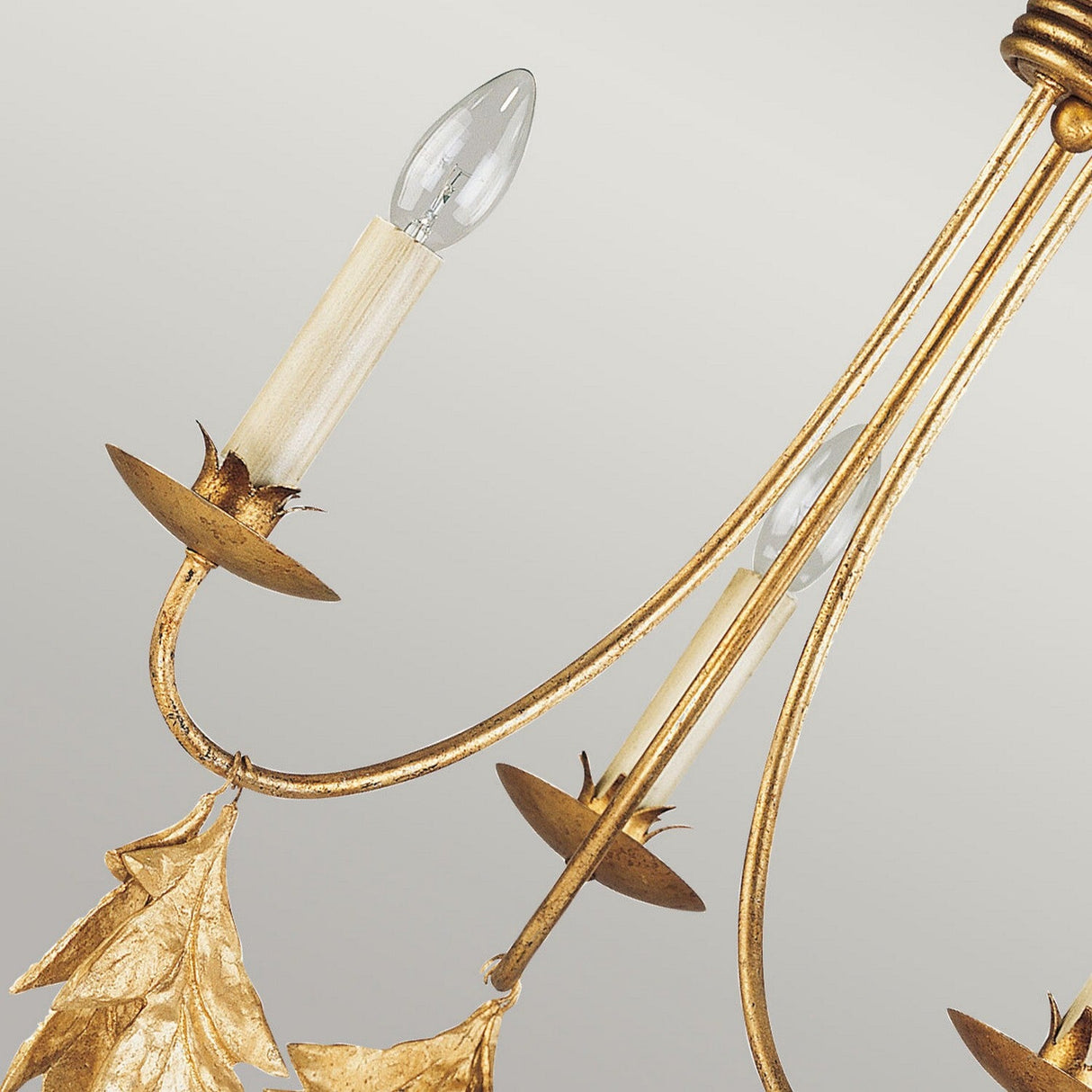 A detailed view of the Sweet Olive 3 Light Chandelier in Gilded Gold reveals its sophisticated design, featuring two candle-shaped light bulbs on gracefully arched, gold-finished branches. The chandelier is adorned with intricate leaf motifs and sparkling crystal medallions, all set against a soft gray backdrop.