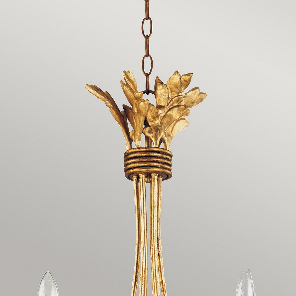 The Sweet Olive 6 Light Chandelier - Gilded Gold features a gleaming gold finish, embellished with leaf-like details at the top and gracefully encircled by a circular band. It is suspended by a delicate chain against a simple gray backdrop.