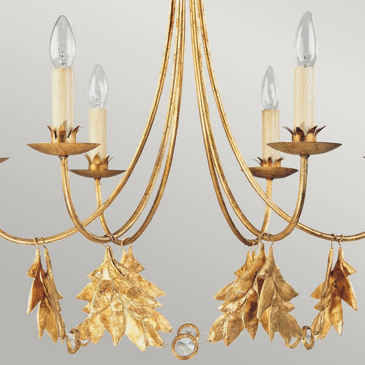 The Sweet Olive 6 Light Chandelier in Gilded Gold showcases a stunning gilded finish, featuring six elegantly curved arms with candle-shaped lights adorned with leaf-like details. The bulbs resemble candle flames, adding a classic and ornate touch against the neutral background.