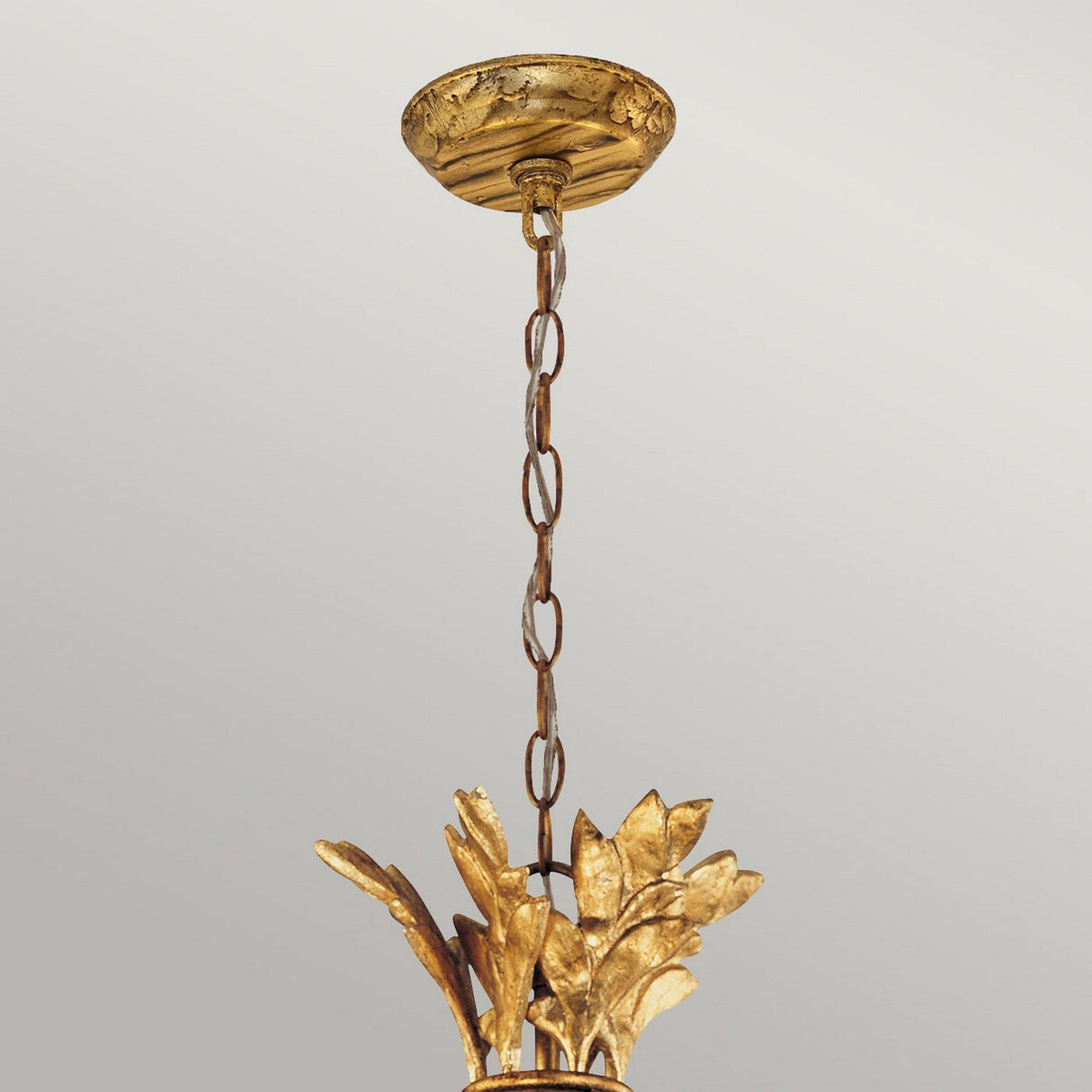 The Sweet Olive 6 Light Chandelier in Gilded Gold showcases leaf-like accents and is suspended from a ceiling mount. The image highlights the top section, featuring a chain that connects the fixture to the ceiling.