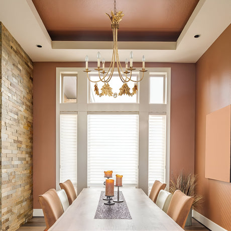 The dining room showcases a contemporary table for six, complete with white and brown chairs. A decorative table runner and candles add charm, while the Sweet Olive 6 Light Chandelier in Gilded Gold gracefully hangs from the tray ceiling. Soft light streams through the blinds, accentuating the earthy tones and stone finishes.