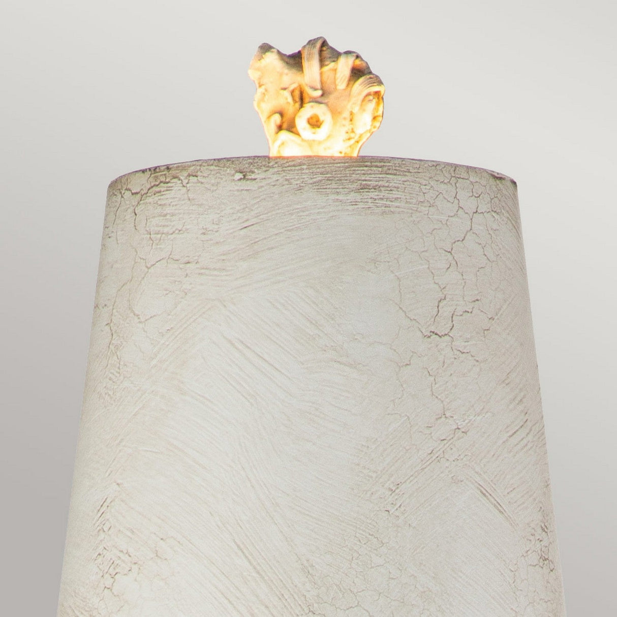 A textured, cylindrical beige object with a cracked finish stands against a light gray background. Atop the cylinder is an antique white shade glowing softly, resembling a small rock or fossil. This elegant table lamp, known as the Swirl 1 Light Table Lamp in White & Black, beautifully blends contemporary design with classic charm.