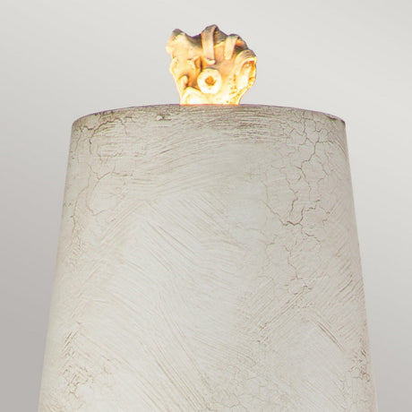 A textured, cylindrical beige object with a cracked finish stands against a light gray background. Atop the cylinder is an antique white shade glowing softly, resembling a small rock or fossil. This elegant table lamp, known as the Swirl 1 Light Table Lamp in White & Black, beautifully blends contemporary design with classic charm.