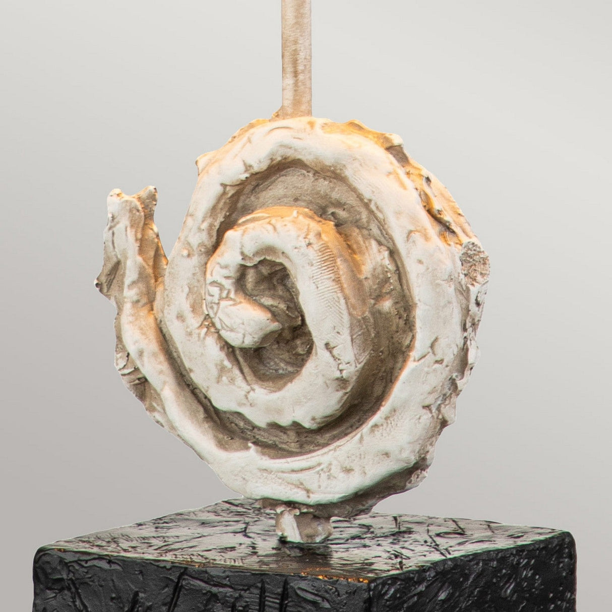 The Swirl 1 Light Table Lamp in White & Black features a spiral-shaped design with a rugged texture, reminiscent of a coiled form, set atop a dark rectangular base. Its weathered, earthy look in shades of white and beige captures the sophisticated elegance of contemporary artistry against a plain background.