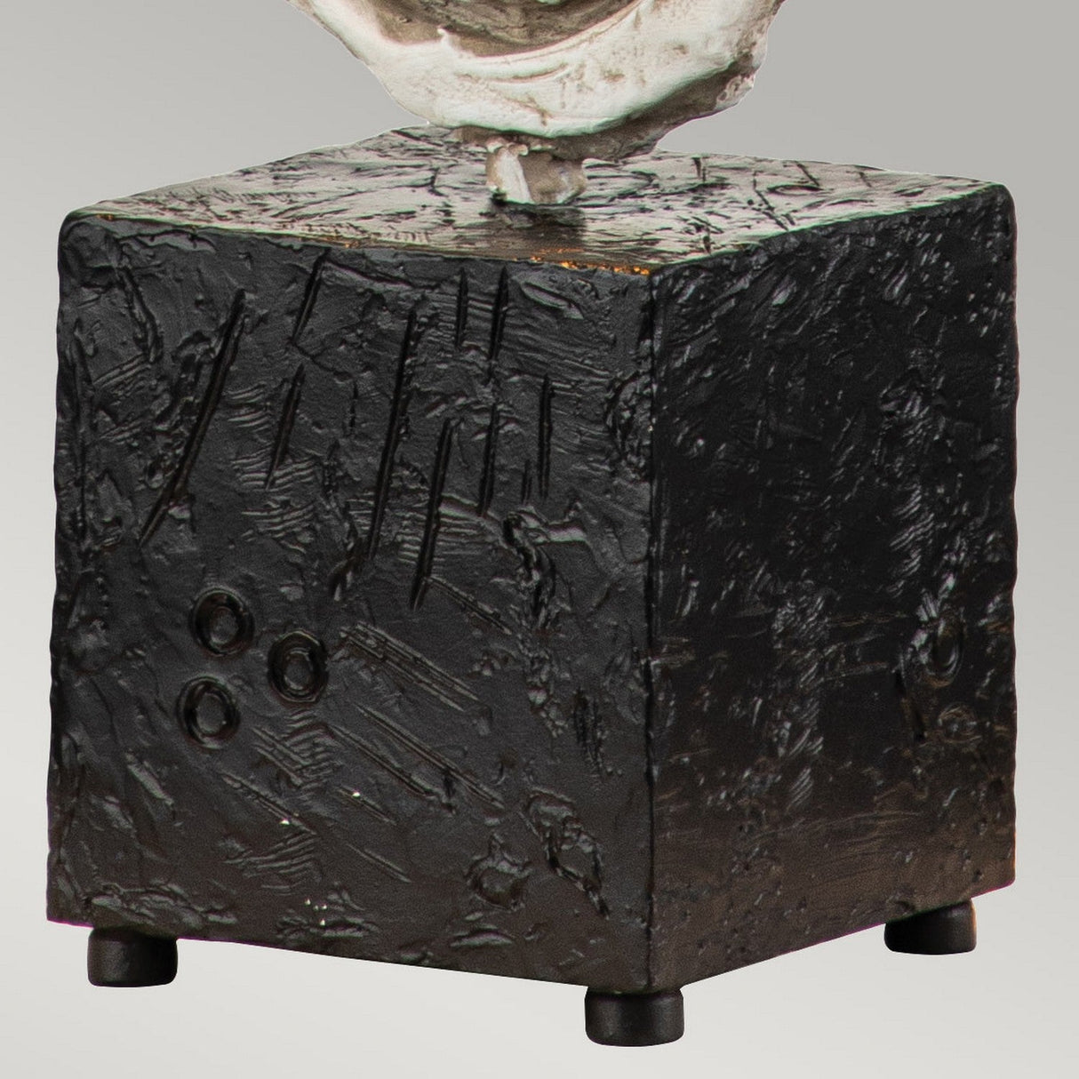 A close-up of the Swirl 1 Light Table Lamp in white and black showcases its textured, dark-colored cube-shaped base with abstract markings and three small circular indentations. This lamp, featuring an antique white shade, stands on four small round feet and embodies a contemporary design reminiscent of modern sculptural elements.