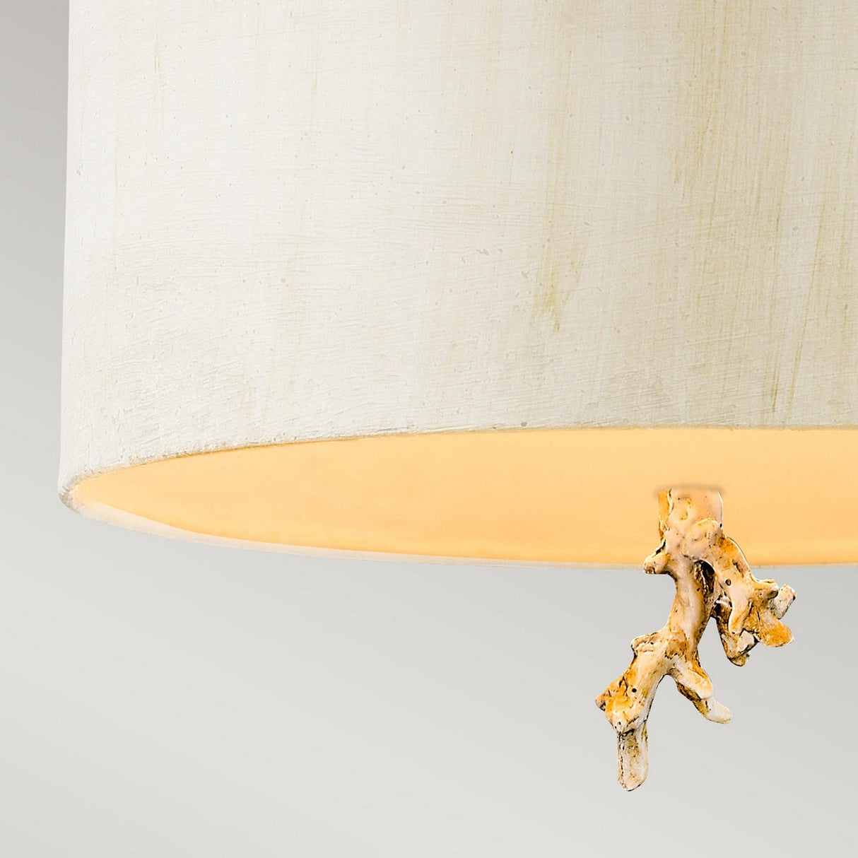 A close-up of the Trellis 1 Light Pendant in Putty Patina and Silver Leaf, featuring a beige fabric shade that emits a warm glow, complemented by an artfully structured beige and orange accent dangling below. The background is a simple, light gray.