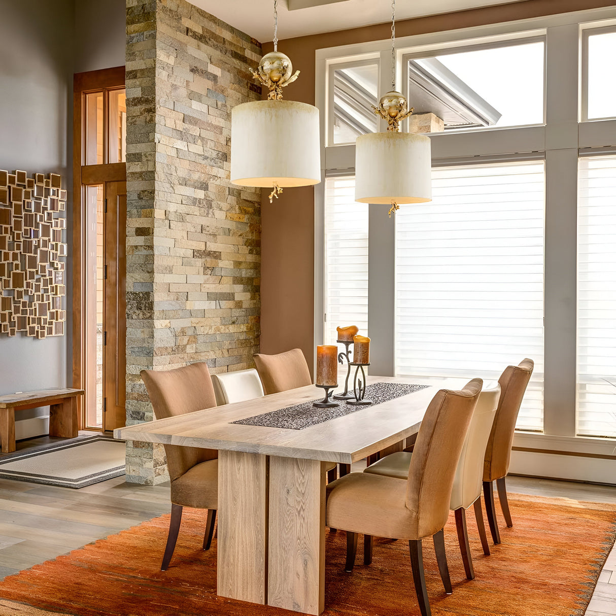 In a contemporary dining room, a wooden table is paired with six beige chairs on an orange rug. Elegantly hanging overhead are two Trellis 1 Light Pendants in Putty Patina & Silver Leaf. A textured stone wall and expansive windows with white blinds brighten the space, further accentuated by a candle centerpiece and captivating wall art.