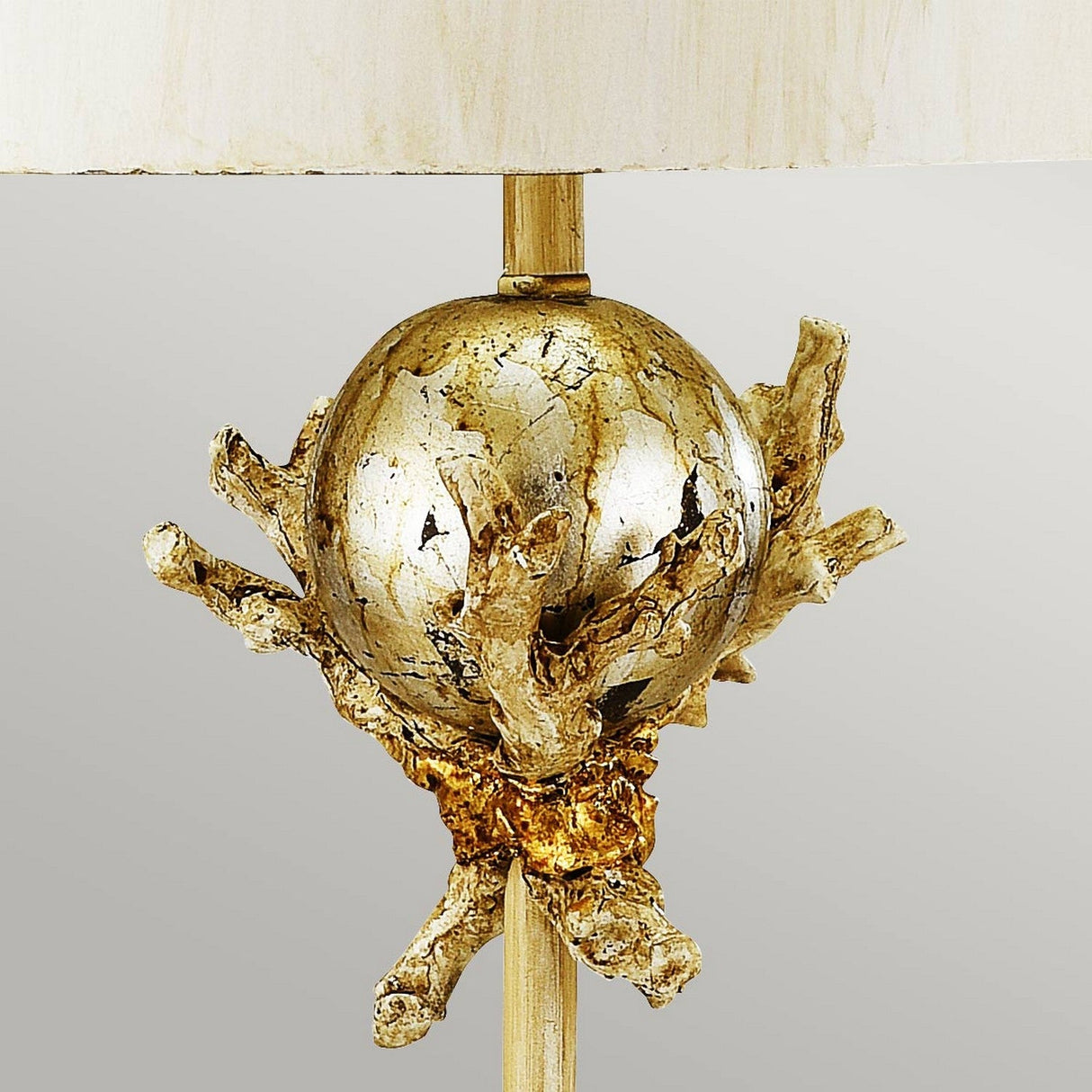 The Trellis 1 Light Table Lamp, finished in Putty Patina & Silver Leaf, features a sophisticated design with a metallic, textured orb base embellished with branch-like extensions. The handcrafted creation includes a neutral lampshade and a vertical post for support, making it an artistic and organic statement lighting piece.