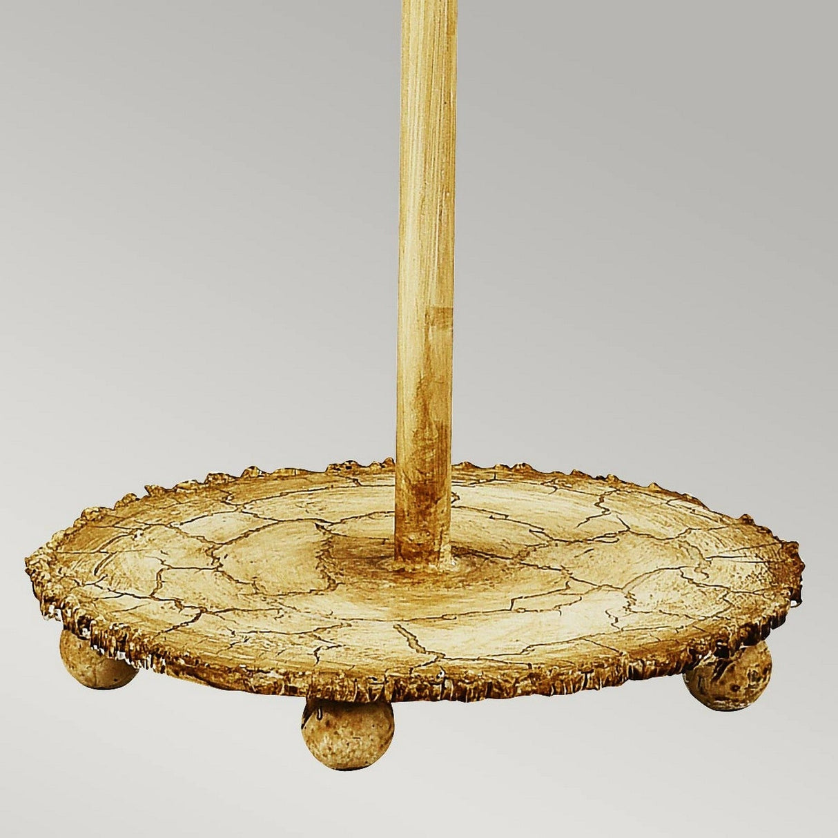 Close-up of the Trellis 1 Light Table Lamp with a round, textured base reminiscent of a tree slice. This handcrafted design showcases natural grain patterns and features four small round legs that support a vertical pole. The graceful lamp, finished in Putty Patina & Silver Leaf, stands out as an eye-catching lighting piece against the plain gray background.