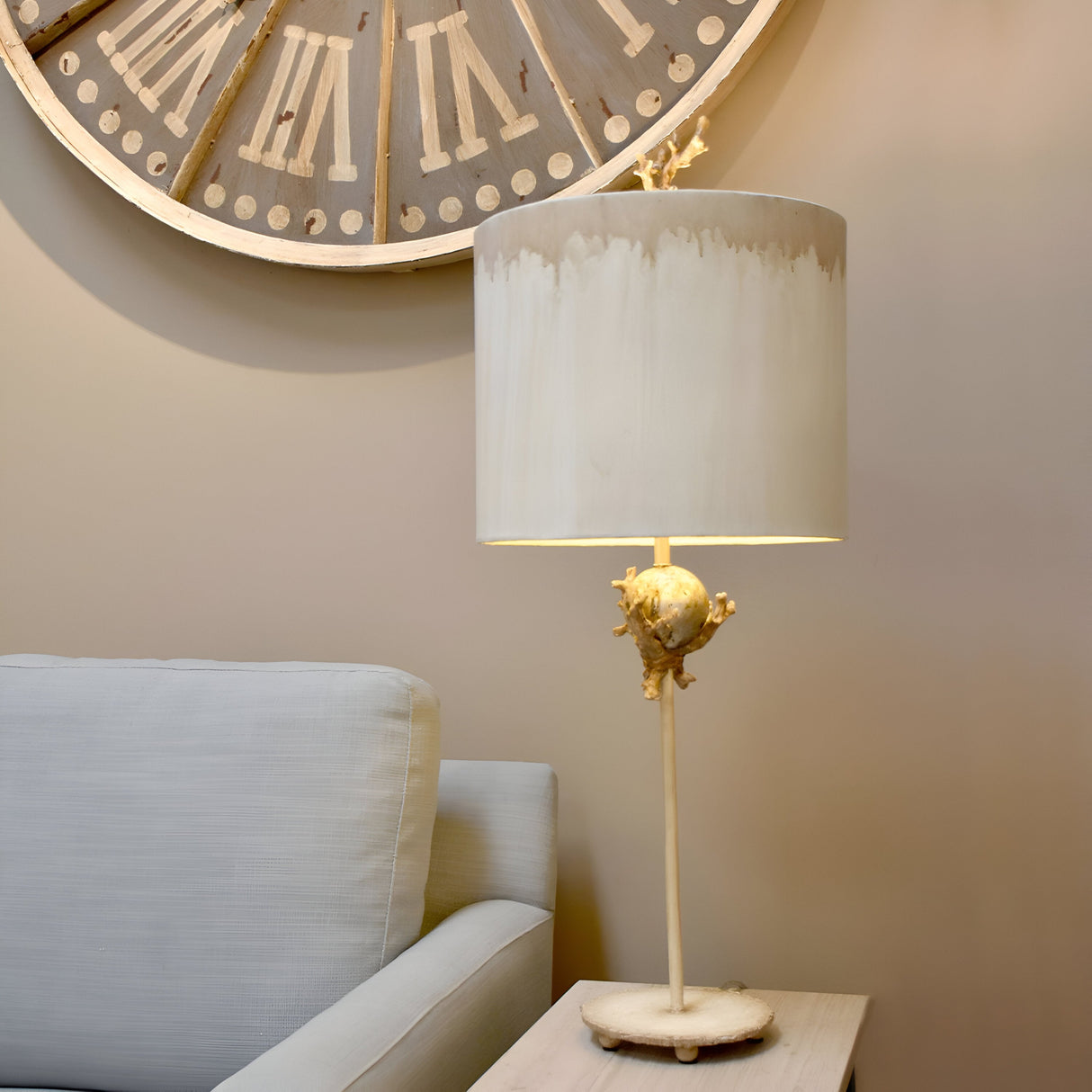 A cozy interior features a white cushioned sofa next to a wooden side table with the Trellis 1 Light Table Lamp in Putty Patina & Silver Leaf. This lamp, adorned with a white shade and unique base, adds charm to the room. A large clock with Roman numerals hangs on the beige wall above.