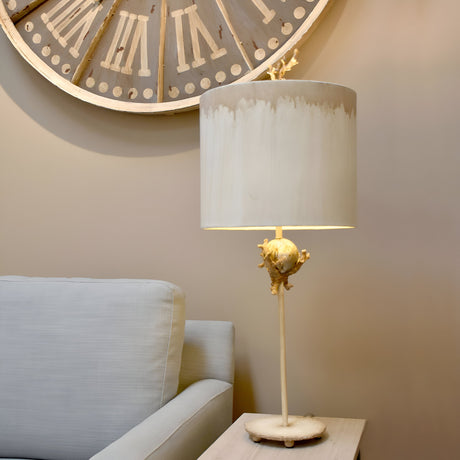 A cozy interior features a white cushioned sofa next to a wooden side table with the Trellis 1 Light Table Lamp in Putty Patina & Silver Leaf. This lamp, adorned with a white shade and unique base, adds charm to the room. A large clock with Roman numerals hangs on the beige wall above.