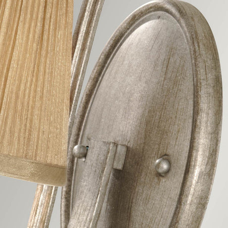 Close-up of the Abbey 1 Light Wall Light in Silver Sand, showcasing its textured metallic design with a ribbed, beige fabric shade. The oval metallic backplate features a Silver Sand finish with subtle weathered detailing, imparting a vintage charm to the design.