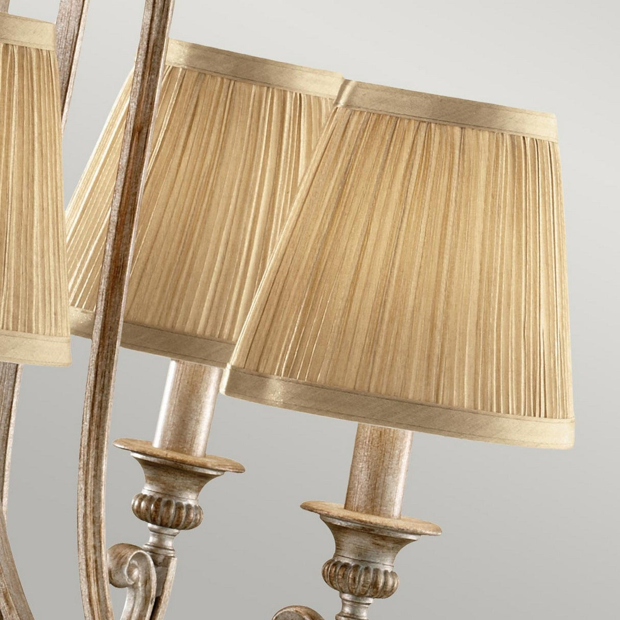 A close-up showcases two elegant table lamps adorned with pleated gold shades. These lamps, featuring ornate vintage-style bases in a Silver Sand finish, are reflected in a mirror, adding to the symmetrical effect against a neutral backdrop. Adding to this sophisticated scene is the Abbey 5 Light Chandelier in Silver Sand.