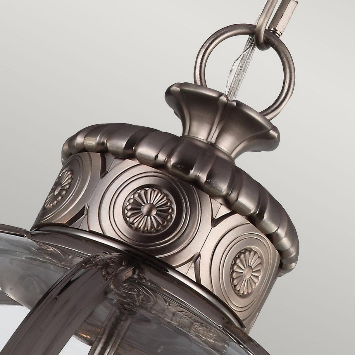 A detailed view of the Adams 4 Light Chandelier - Antique Nickel highlights its Victorian-inspired elegance, featuring intricate circular patterns and a metallic finish. The glass element harmonizes beautifully with the Antique Nickel chain against a simple background.