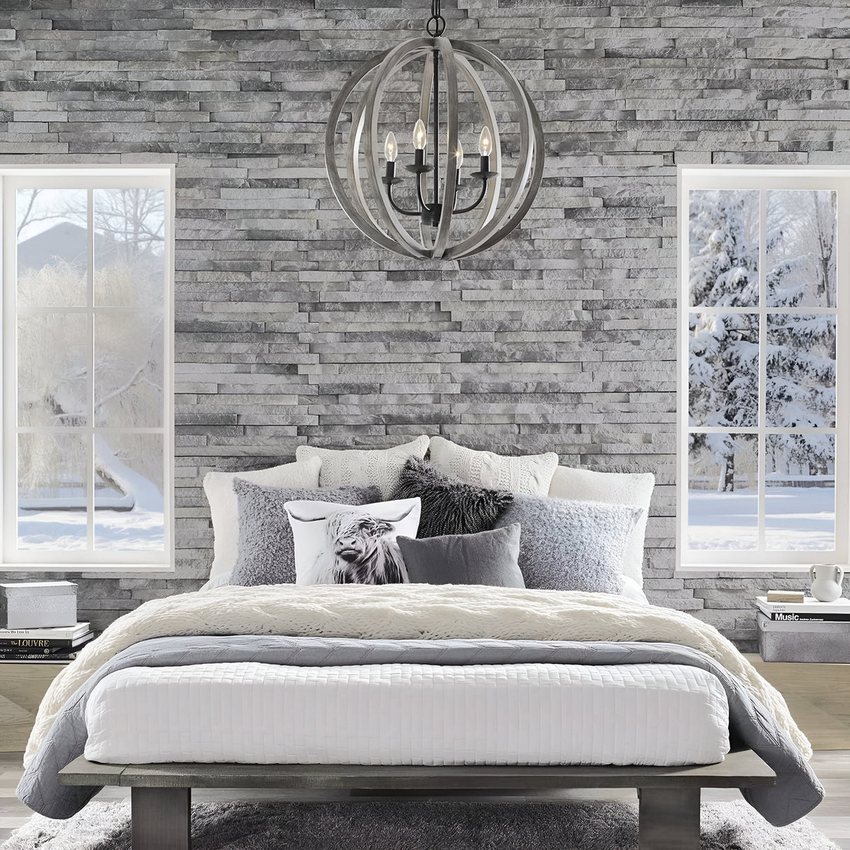A cozy bedroom with a gray stone accent wall showcases the Allier 4 Light Chandelier in weathered oak wood and antique forged iron above. The bed is adorned with layers of white and gray bedding complemented by decorative pillows, while large windows offer a view of the snowy winter landscape outside.