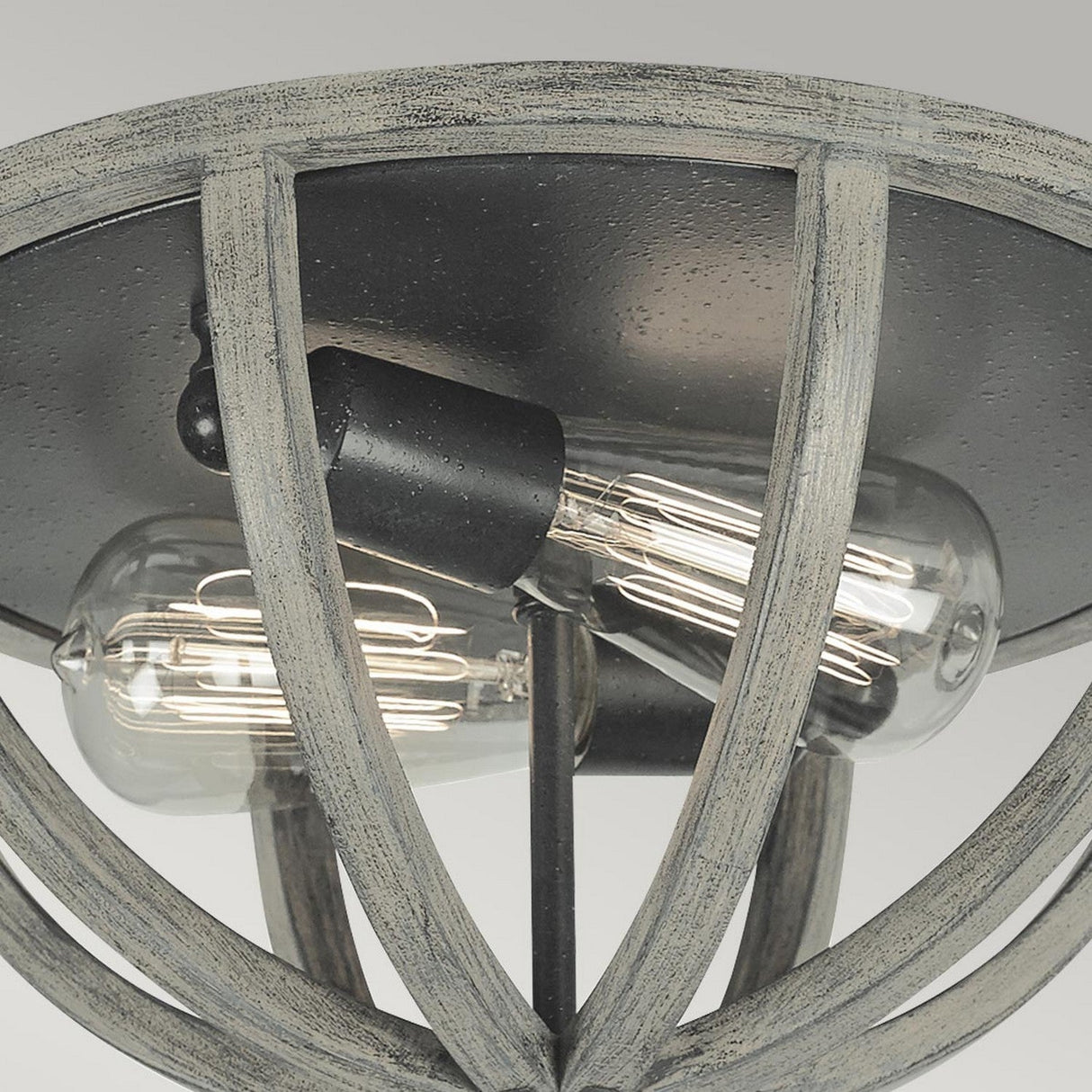 A close-up of the Allier 2 Light Flush showcases a rustic light fixture with two vintage-style exposed bulbs set in an antique forged iron frame. This design seamlessly blends industrial and antique elements with a textured finish that accentuates the visible filaments.