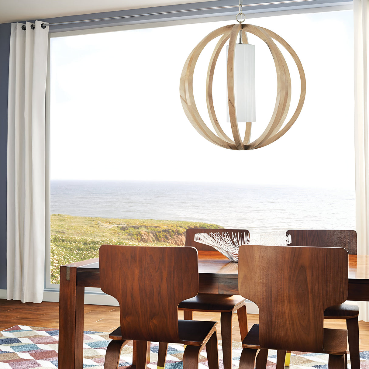 A modern dining room with a large window view of the ocean features a wooden table accompanied by four matching chairs, all with a light wood finish. The space is illuminated by the Allier 1 Light Large Pendant in light wood and brushed steel, perfectly complementing its surroundings. White curtains elegantly frame the window, and a checkered rug lays gracefully on the hardwood floor.