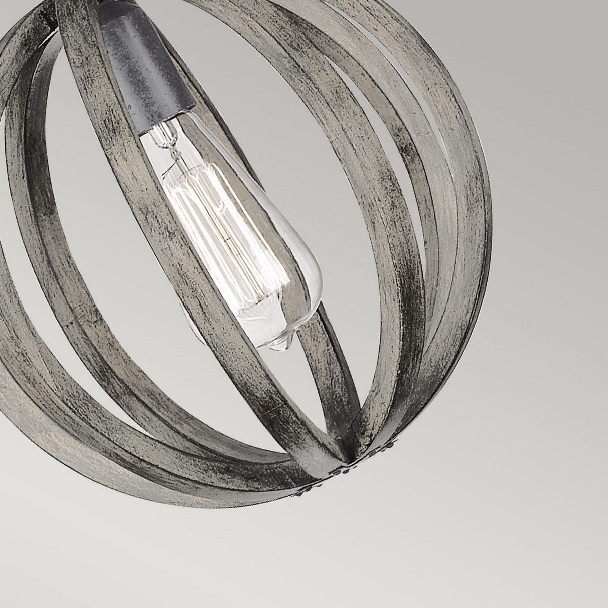 A modern mini pendant light fixture, the Allier 1 Light Mini Pendant, showcases a round cage crafted from curved metal bands around an exposed vintage-style bulb. The metal features an antique forged iron finish, complemented by a weathered oak wood element against a soft, neutral gray background.