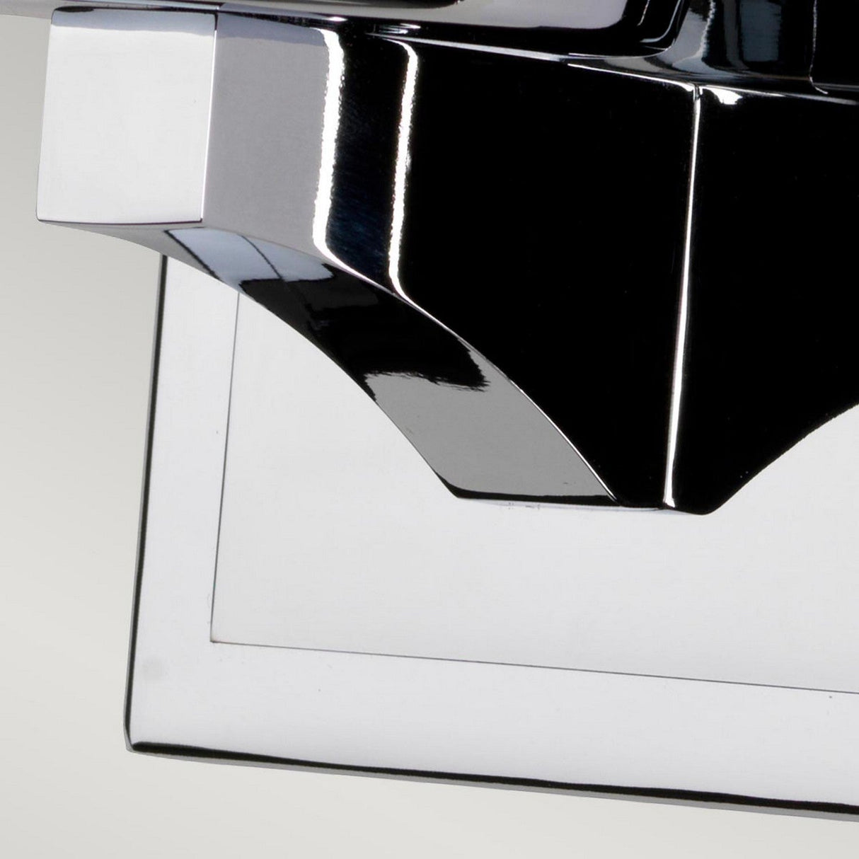 Close-up of a shiny metallic object with angular edges, featuring reflective surfaces and a geometric design reminiscent of the Amalia 1 Light Large Wall Light - Polished Chrome. The background is a light gray, emphasizing the object's sleek and modern appearance, enhanced by its IP44 rating for durability.