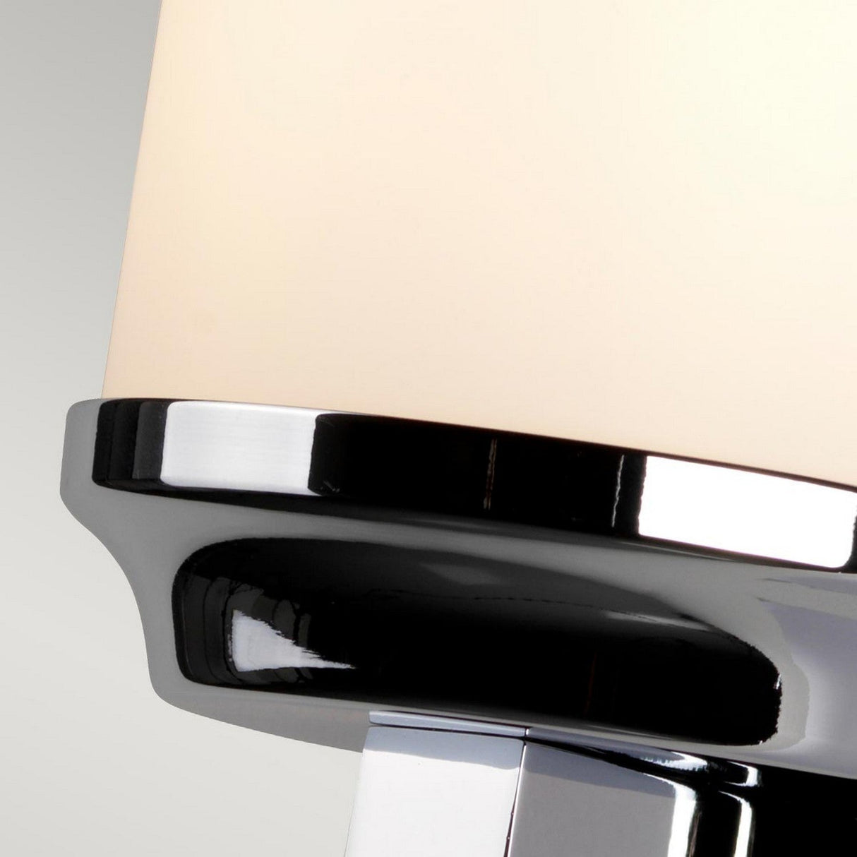 A close-up of the Amalia 1 Light Small Wall Light showcases its modern lampshade, featuring a smooth white surface and a shiny polished chrome rim. Its sleek and minimalist design is ideal for bathroom lighting, set against a light gray background.
