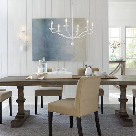 A dining room exuding modern sophistication features a wooden table and six beige chairs, with the Annie 6 Light Chandelier in Textured Plaster White elegantly hanging above. The table is set with glasses, a water carafe, and dishes. A large abstract painting and a wall sconce adorn the white-paneled wall as natural light filters through the window.