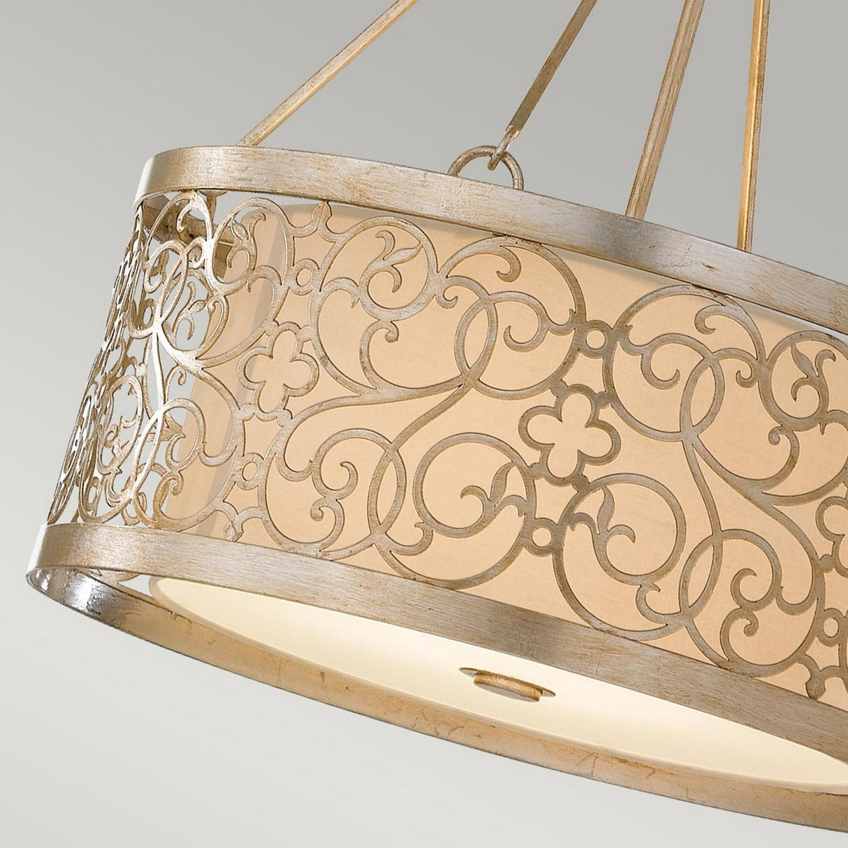The Arabesque 4 Light Chandelier, complete with a Silver Leaf Patina finish, boasts an exquisite metal grille design that adds a luxurious touch. Its intricate pattern elegantly casts enchanting shadows on the milky glass diffuser, while it gracefully hangs from three cords against a soft light gray background.