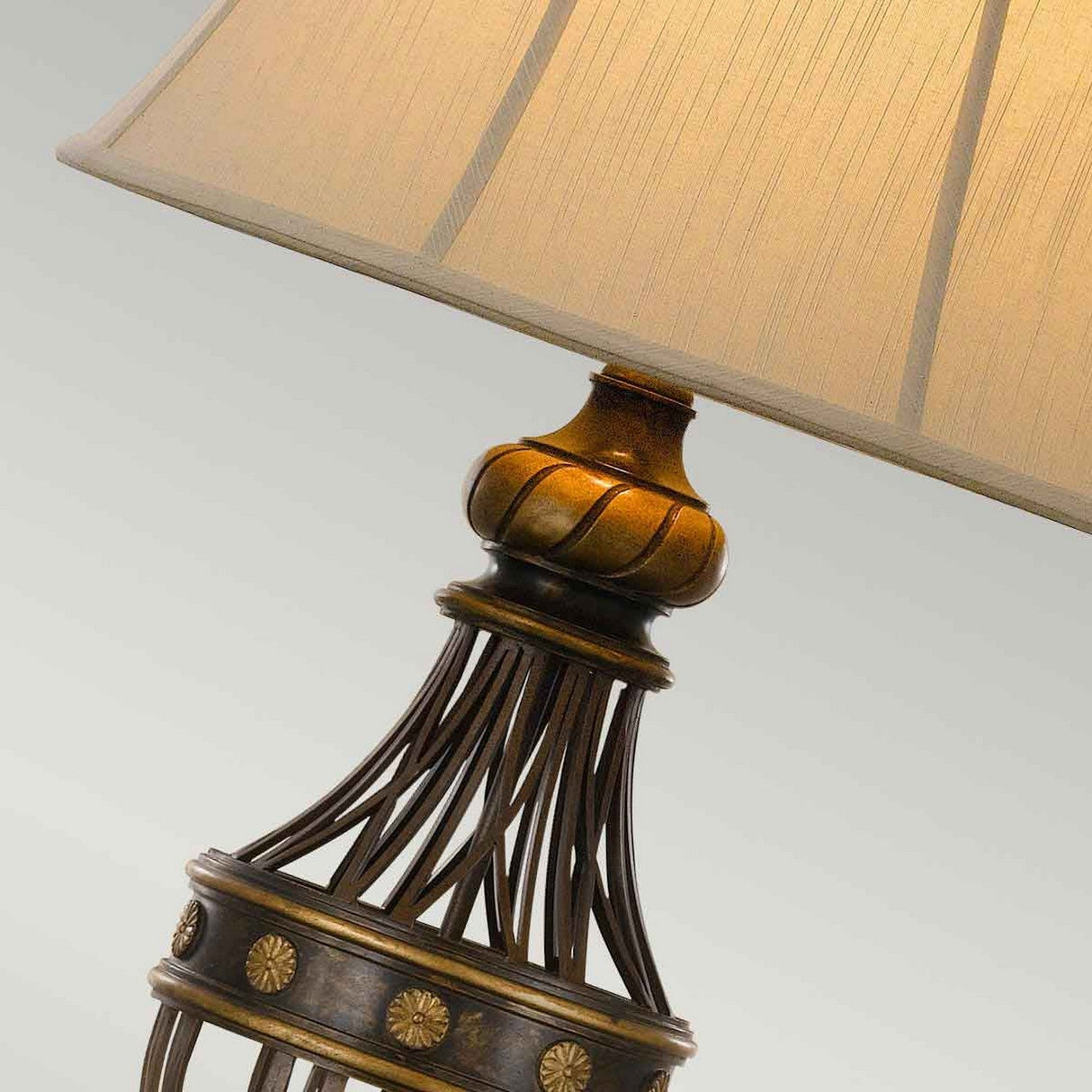 Close-up of the Augustine 1 Light Table Lamp in Antique Brown with a beige fabric lampshade. The base showcases an ornate bronze design with vertical bars and circular motifs, ideal for Classic Design in Transitional Interiors. The background is a plain, neutral color.