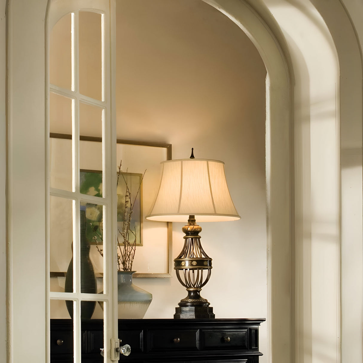 In a cozy interior, the Augustine 1 Light Table Lamp in Antique Brown sits atop a dark wooden table, encircled by vases and framed artwork. The view through the glass-paneled door allows soft shadows to dance on the walls, perfectly embodying the warmth of transitional interiors.