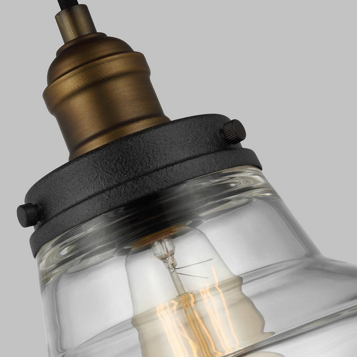 Close-up of the Baskin-1 Light Pendant featuring a painted aged brass and dark weathered zinc finish. The clear shade beautifully reveals the bulb inside, casting a warm glow that adds rustic charm. A neutral gray background enhances its industrial-inspired design.