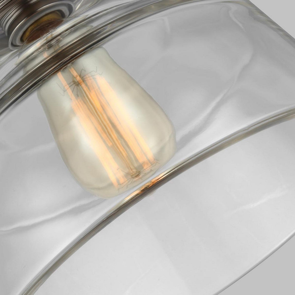 Close-up of a clear glass pendant light featuring an exposed filament bulb. The glowing filaments are visible inside, with the fixture's polished nickel details subtly appearing at the top. The background is neutral.