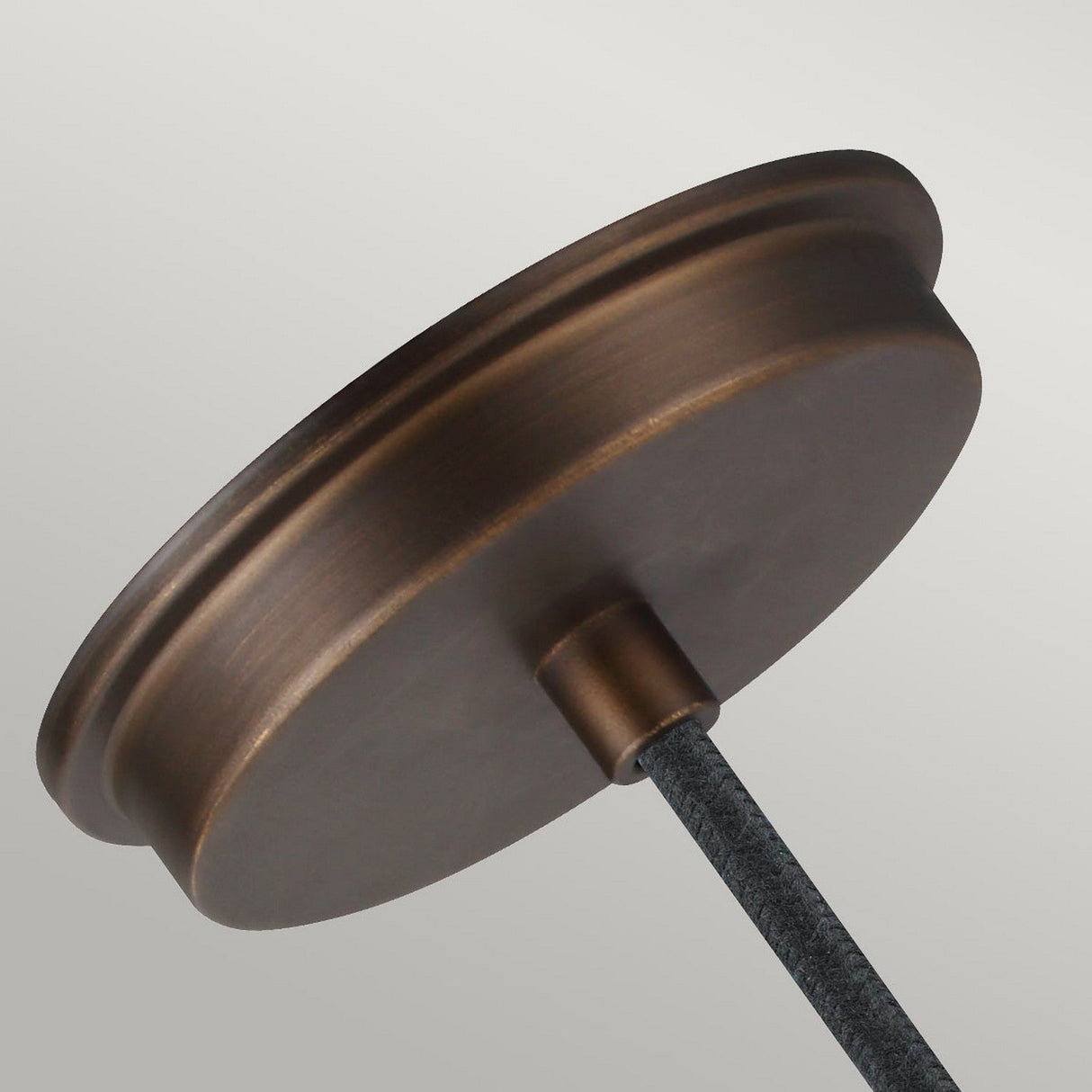 Close-up of a round, aged brass canopy from the Baskin-2 1 Light Pendant in Painted Aged Brass/Dark Weathered Zinc, featuring a textured, dark-colored cord extending downwards. This pendant light's canopy has a smooth, slightly reflective surface and is set against a plain, light gray background.