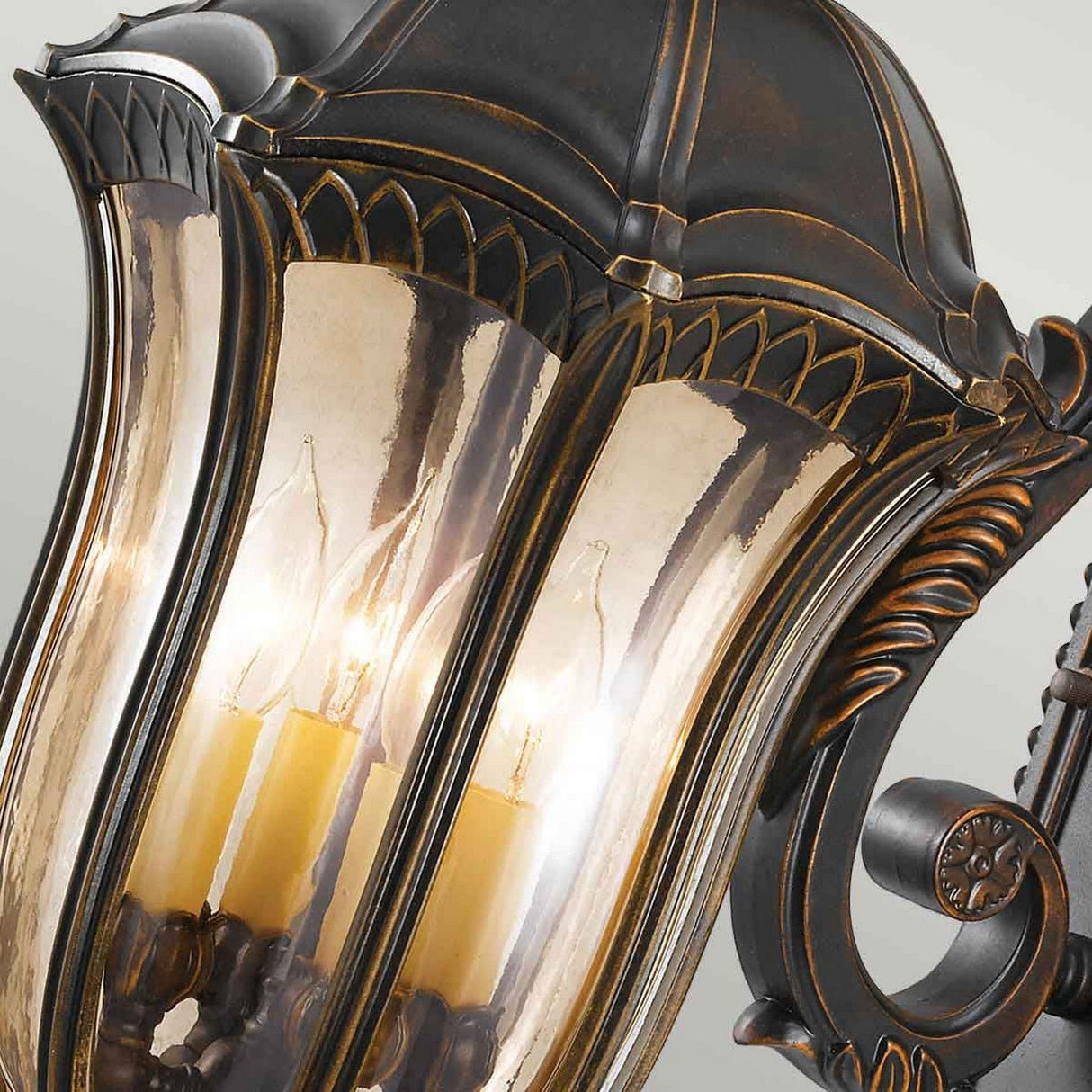 The Baton Rouge 4 Light Outdoor Large Wall Lantern in Walnut showcases a close-up of a decorative black and bronze design with intricate detailing and transparent panels, revealing four candle-shaped bulbs glowing inside. This vintage and elegant large wall light is ideal for outdoor lighting, bringing a sophisticated touch with its subtle walnut finish accents.