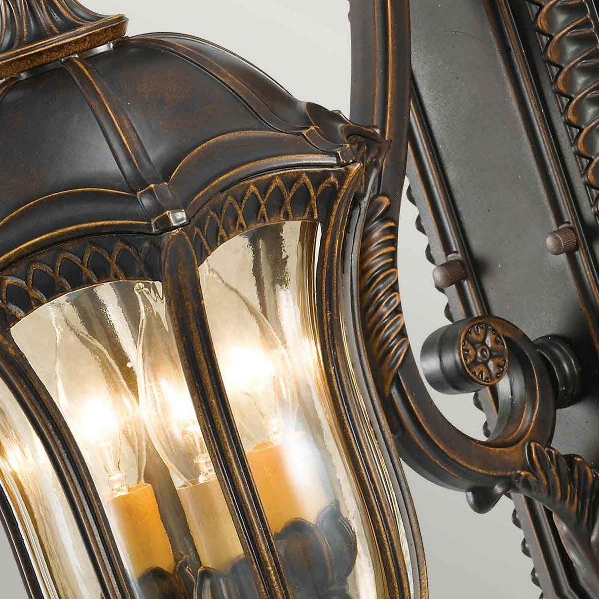 Close-up of the Baton Rouge 3 Light Outdoor Medium Wall Lantern - Walnut, showcasing its ornate design with intricate detailing. The lantern, exuding Baton Rouge charm, features three illuminated bulbs encased in clear glass, offering a vintage, weathered finish that enhances its classic and elegant appeal.
