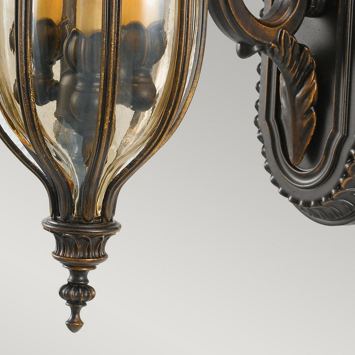 A close-up of the Baton Rouge 3 Light Outdoor Medium Wall Lantern in walnut showcases its ornate carved metal design and three enclosed candles. The aged bronze finish exudes classic elegance, reminiscent of Baton Rouge charm.