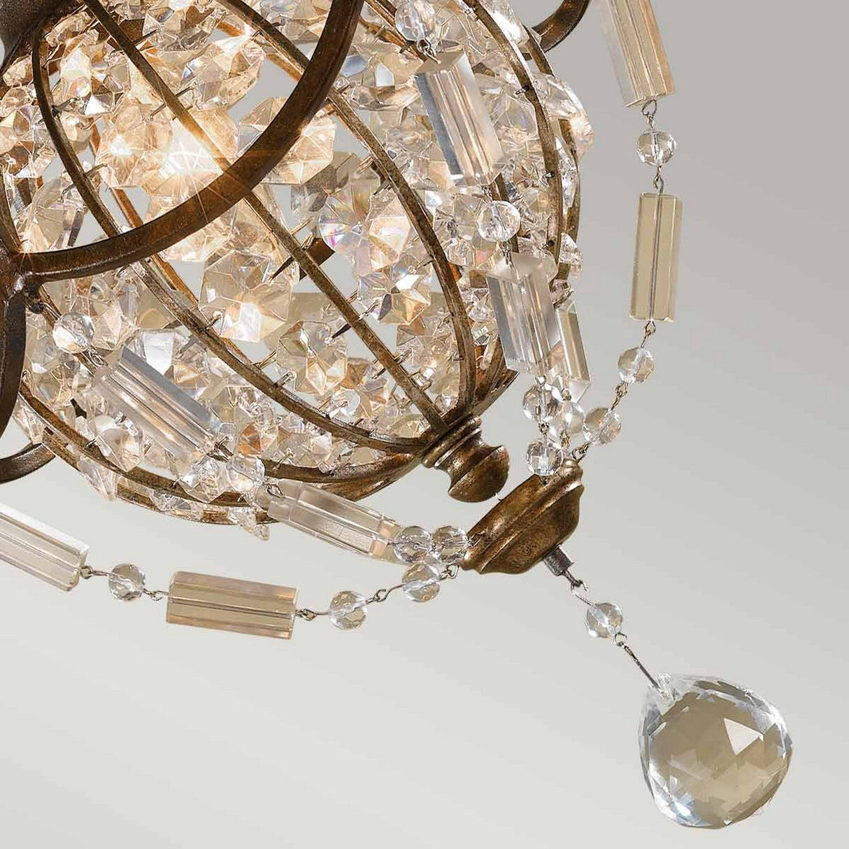 A close-up of the Bellini 1 Light Pendant Light reveals its spherical metal frame, adorned with antique quartz crystals and beads. A large, clear crystal at the bottom sparkles under soft lighting. The oxidized bronze finish enhances its vintage charm against a gradient gray background.