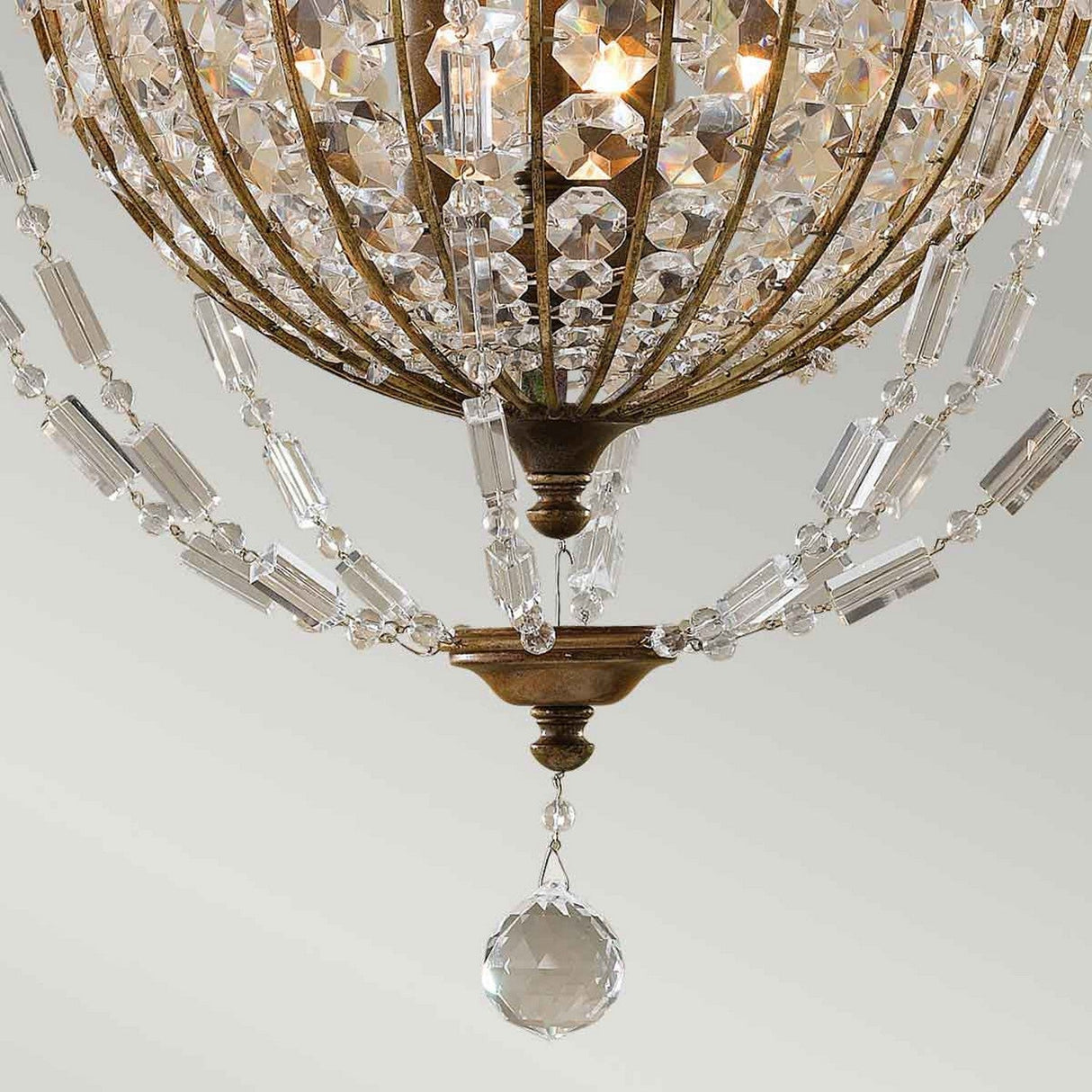 A close-up of the Bellini 3 Light Chandelier, showcasing its elegant antique quartz crystal bead strands and large crystal drop. The chandelier's oxidized bronze metallic frame, reminiscent of 1920s French Chateau style, features intricate detailing and is softly illuminated from within.