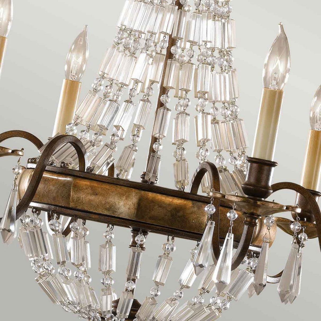 A close-up of the Bellini 6 Light Chandelier showcases candle-shaped bulbs and cascading antique quartz crystals on an oxidized bronze/British bronze metal frame. The crystals reflect light, creating a shimmering effect against a soft, neutral background.