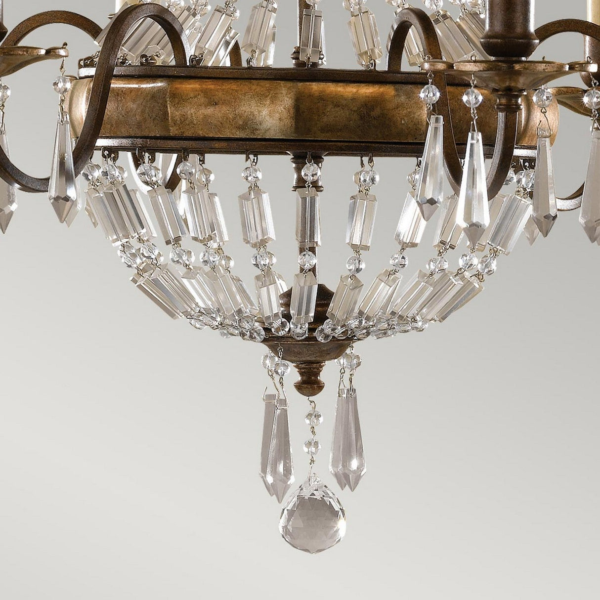A close-up of the Bellini 6 Light Chandelier showcases its luxurious design, featuring an ornate display of clear crystal prisms and pendants gracefully hanging from an oxidized bronze and British bronze circular frame. Antique quartz crystals add a touch of timeless elegance against the plain, light gray background.