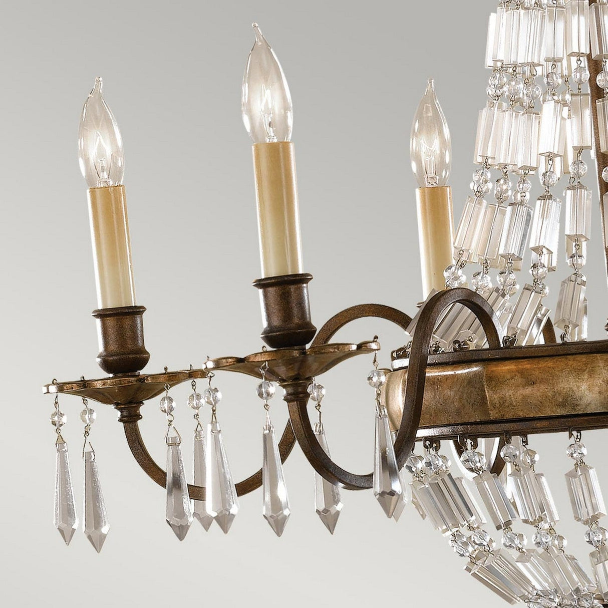 The Bellini 6 Light Chandelier - Oxidized Bronze/British Bronze features a close-up view of its splendid design, showcasing three candle-like light bulbs. It is elegantly crafted with a bronze metal frame and lavishly decorated with numerous antique quartz crystal pendants that beautifully reflect the light, embodying luxury lighting against a soft, neutral background.