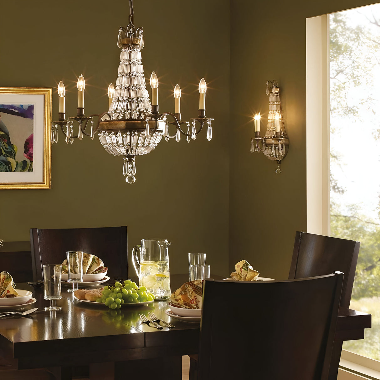 The dining room showcases a dark wood table set for four with plates, glasses, a pitcher of lemon water, and green grapes. Above the table hangs the elegant Bellini 6 Light Chandelier in Oxidized Bronze/British Bronze, casting luxurious lighting over the olive green walls and through the large window that overlooks a garden scene.