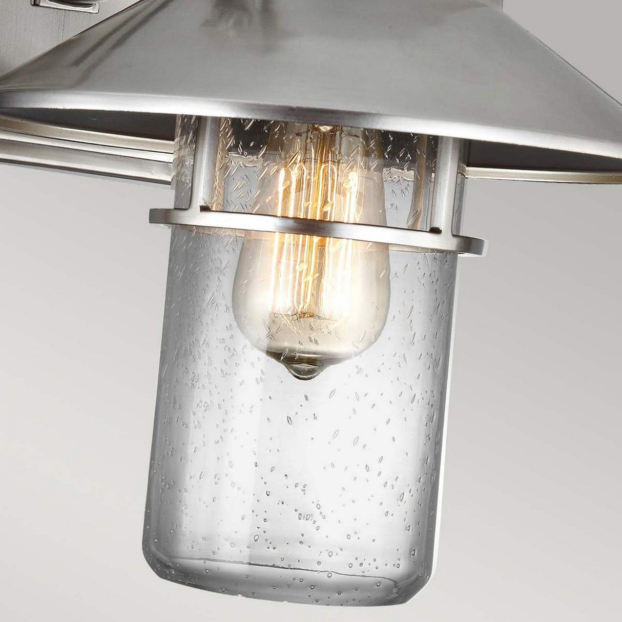 A close-up of the Boynton 1 Light Outdoor Medium Wall Lantern in Painted Brushed Steel showcases a modern metal fixture with a clear glass shade featuring raindrop texture. Inside, a glowing filament bulb emits warm light. The painted brushed steel finish enhances its contemporary appeal, making it perfect for any outdoor setting.