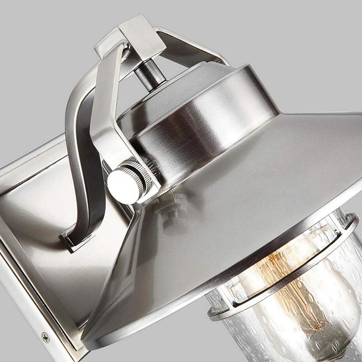 Close-up of the Boynton 1 Light Outdoor Small Wall Lantern in a painted brushed steel finish. This weather-resistant fixture showcases a clear glass casing around an exposed bulb, highlighted by its sleek silver mount and bracket set against a gray backdrop, making it ideal for outdoor environments.