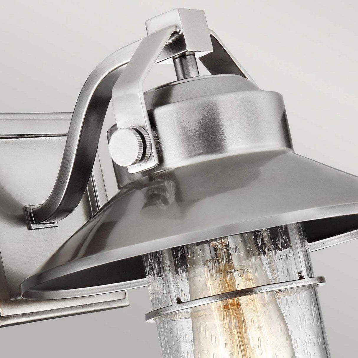 Close-up of the Boynton 1 Light Outdoor Small Wall Lantern, featuring a painted brushed steel finish and clear seeded glass shade that reveals the bulb inside. This weather-resistant fixture showcases a sleek, industrial design and is mounted on a matching square base.