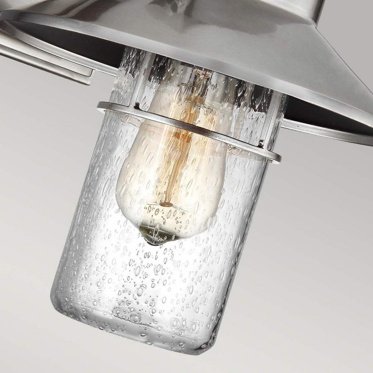 Close-up of the Boynton 1 Light Outdoor Small Wall Lantern in a painted brushed steel finish, highlighting a glowing vintage-style bulb encased in a clear glass shade. The textured, bubbled appearance of the glass brings an artistic flair to its industrial-modern design.