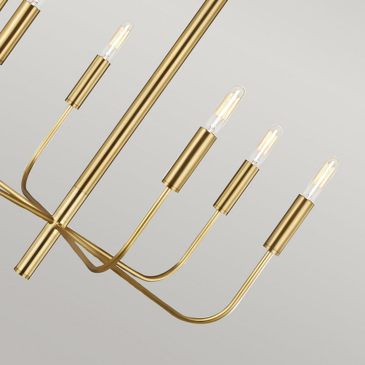 The Brianna 6 Light Chandelier - Burnished Brass features sleek, curved arms that extend outward, each holding a slender tubular bulb. Its design radiates modern elegance with its minimalist style against a simple, light background.